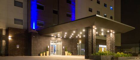 A hotel entrance with a covered walkway, large potted plants, and a blue-lit facade.