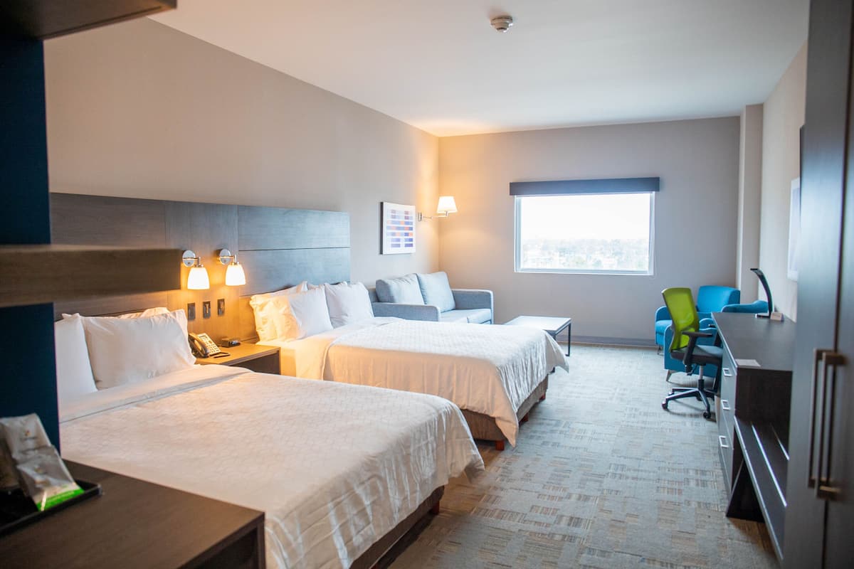 A hotel room with two beds, a desk, a chair, a window, and a wall-mounted lamp.