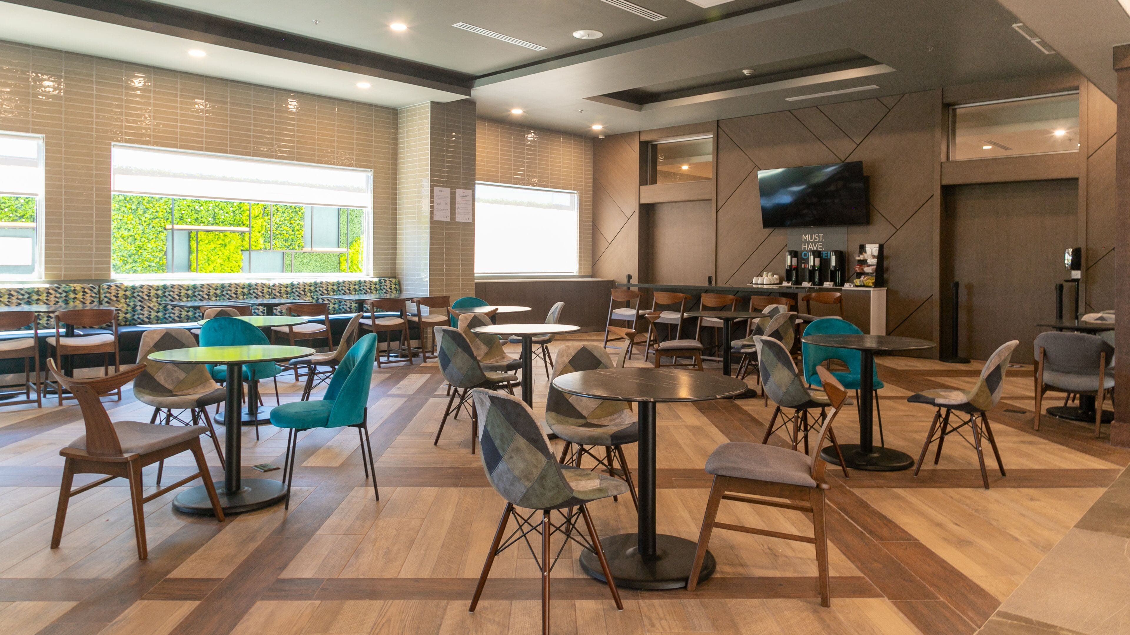 Una cafetería moderna con suelo de madera, diversas opciones de asientos que incluyen mesas y sillas, y un ventanal con vistas a zonas verdes.