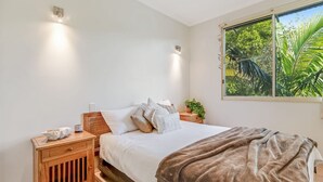 A bedroom with a bed, nightstands, a window with a view of greenery, and wall-mounted lights. - Baden 28 - Rainbow Shores. Top Floor Unit. Air conditioned. Walk to Beach (Rainbow Beach)