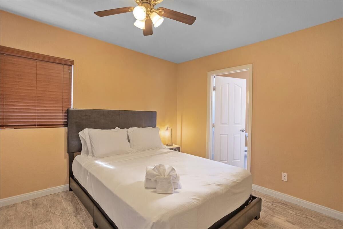 Una cama bien tendida con sábanas blancas y una toalla doblada, un ventilador de techo, una lámpara de noche y una puerta en una habitación de paredes color beige.