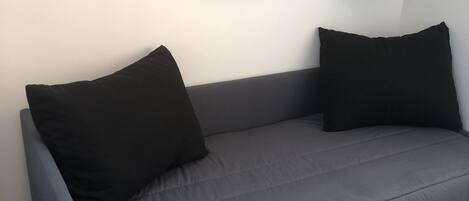 En grå soffa med svarta kuddar, en vit vägg och tre grå hyllor som är tomma.