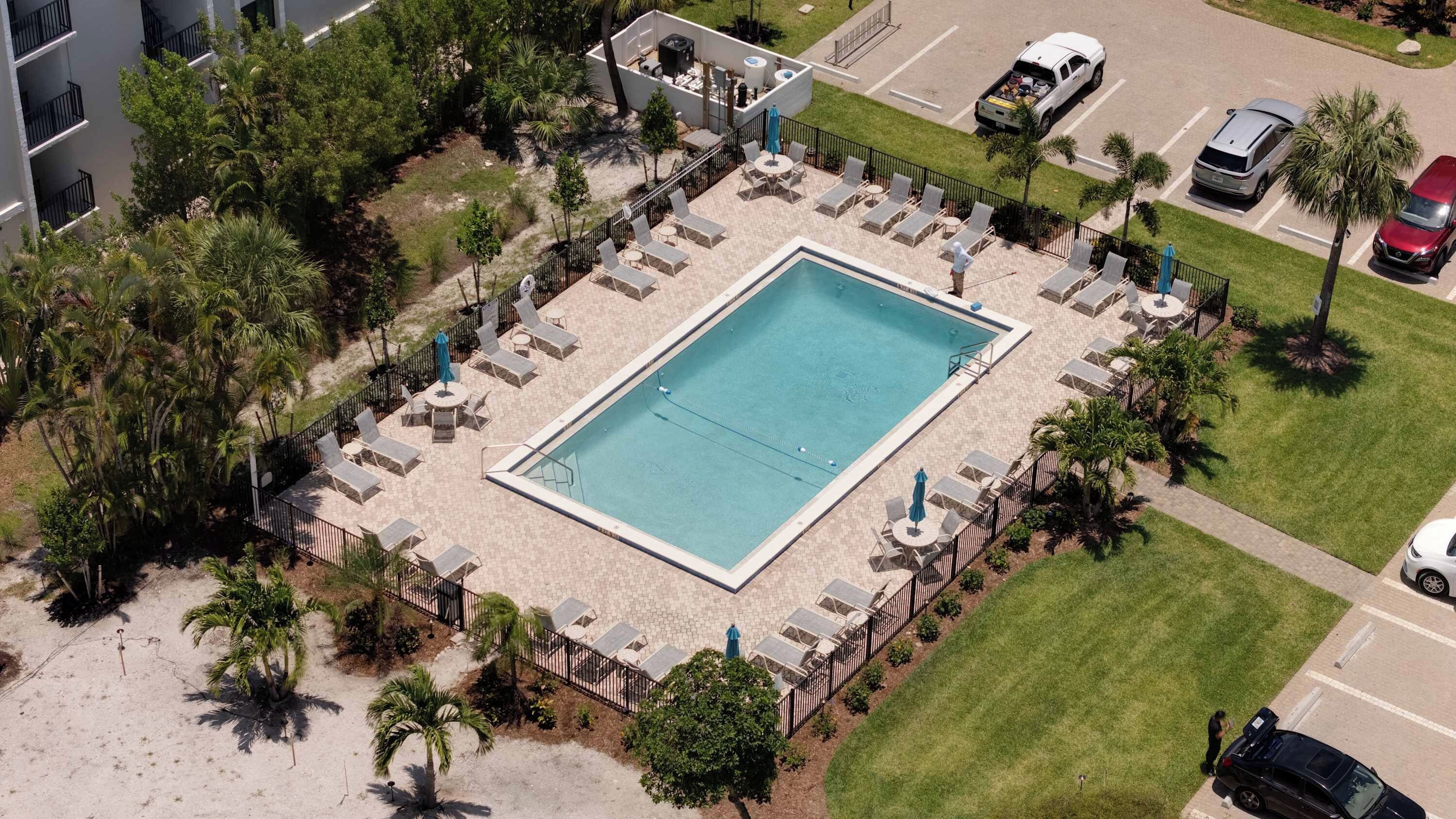 Oversigtsbillede af et hotel med en rektangulær pool omgivet af liggestole og parasoller, et parkeringsområde med biler og en anlagt have med palmer.