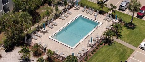 Oversigtsbillede af et hotel med en rektangulær pool omgivet af liggestole og parasoller, et parkeringsområde med biler og en anlagt have med palmer.