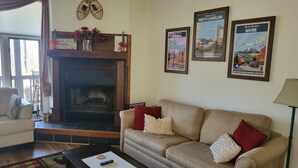 A cozy living room with a fireplace, a sofa, and a coffee table. There are framed posters on the wall and a window with curtains.