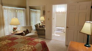 A hotel room with a bed, two chairs, a small table, and a lamp. There is a door leading to another room.
