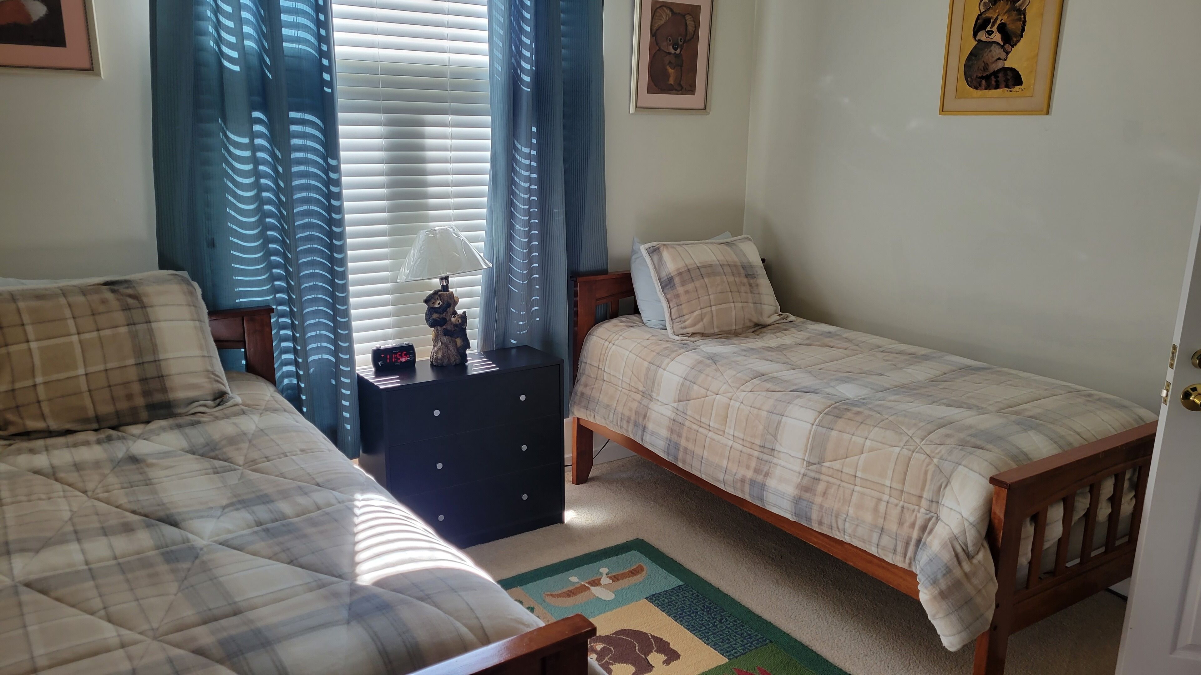A bedroom with two beds, a nightstand with a clock, a lamp, and a teddy bear.