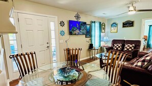 A dining area with a glass table surrounded by wooden chairs, a television mounted on the wall, and a ceiling fan. - Book Fast! Top-Rated 5★ Cottage Near Carolina Beach—Private, Peaceful, Perfect (Wilmington)