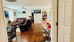 A spacious living room with a dark brown hardwood floor, a brown leather sofa, and a dining area with a glass table and metal chairs. - Book Fast! Top-Rated 5★ Cottage Near Carolina Beach—Private, Peaceful, Perfect (Wilmington)