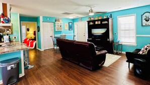A spacious living room with a dark brown leather sofa, a television, and a kitchen area with a marble countertop and red chairs. - Book Fast! Top-Rated 5★ Cottage Near Carolina Beach—Private, Peaceful, Perfect (Wilmington)