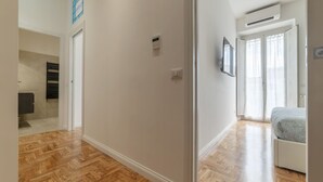 Un couloir doté d’un plancher en bois, une porte donnant sur une pièce avec un lit et une fenêtre, et un appareil de climatisation fixé au mur.
