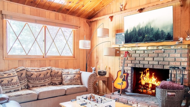 A cozy cabin interior with a sofa, a foosball table, a fireplace, and a television mounted above it. - PRIME Location~Walk to Ski Shuttle ~ Fully Fenced ~ EV Charger ~ Mini Sled Hill (Big Bear Lake)