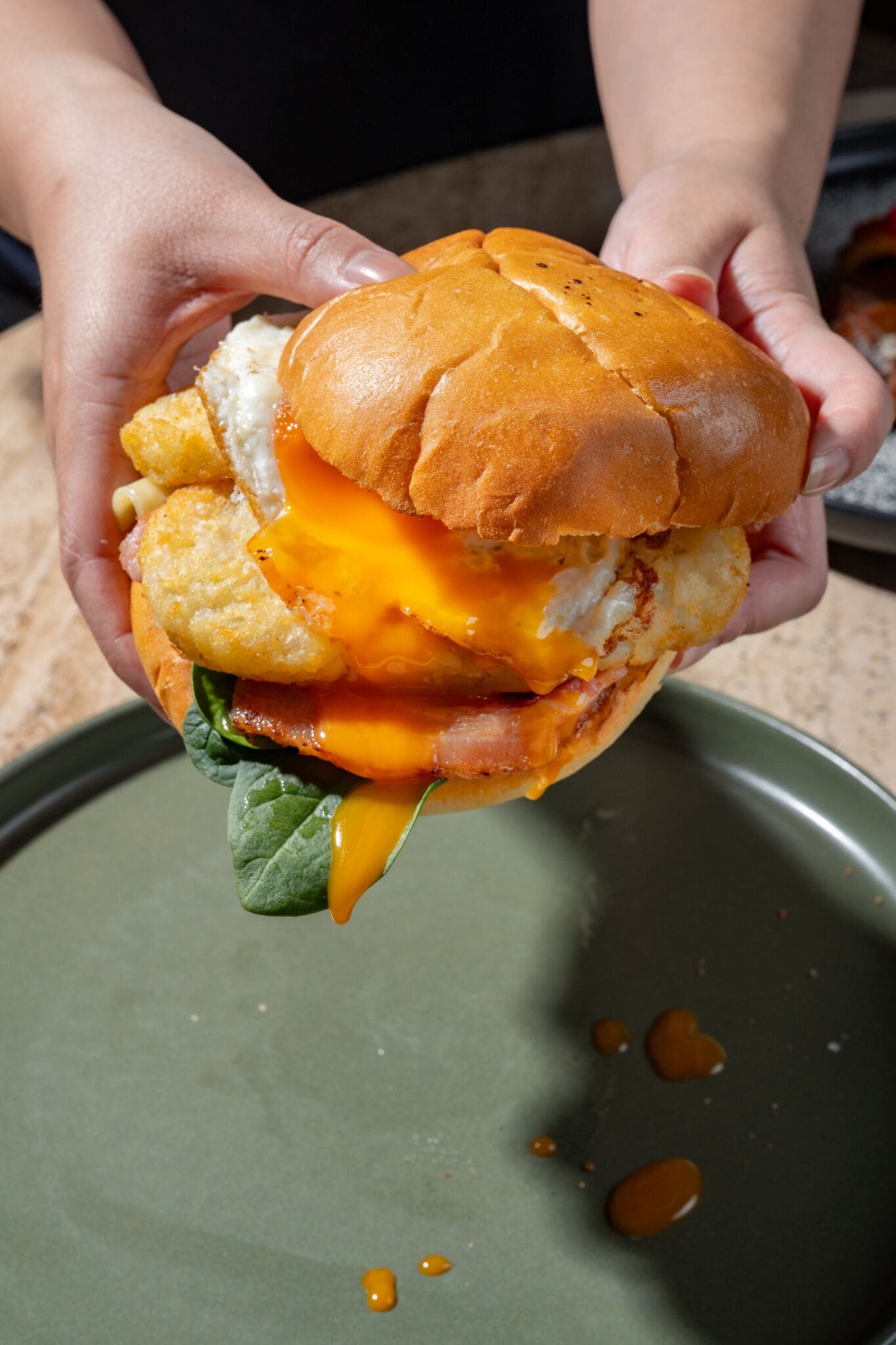 A sandwich held by hands, with visible ingredients like cheese, bacon, and a leaf.