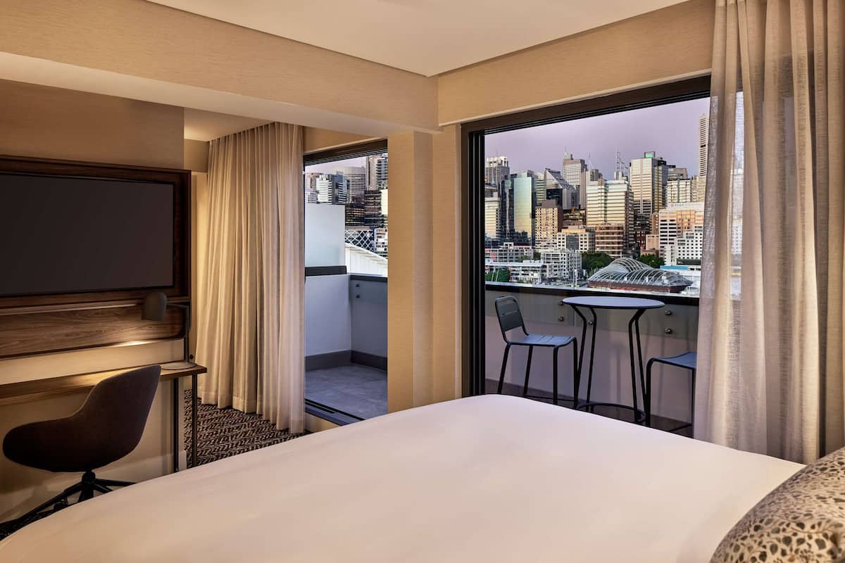 A hotel room with a bed, a TV, a chair, a balcony with a table and chairs, and a city view.