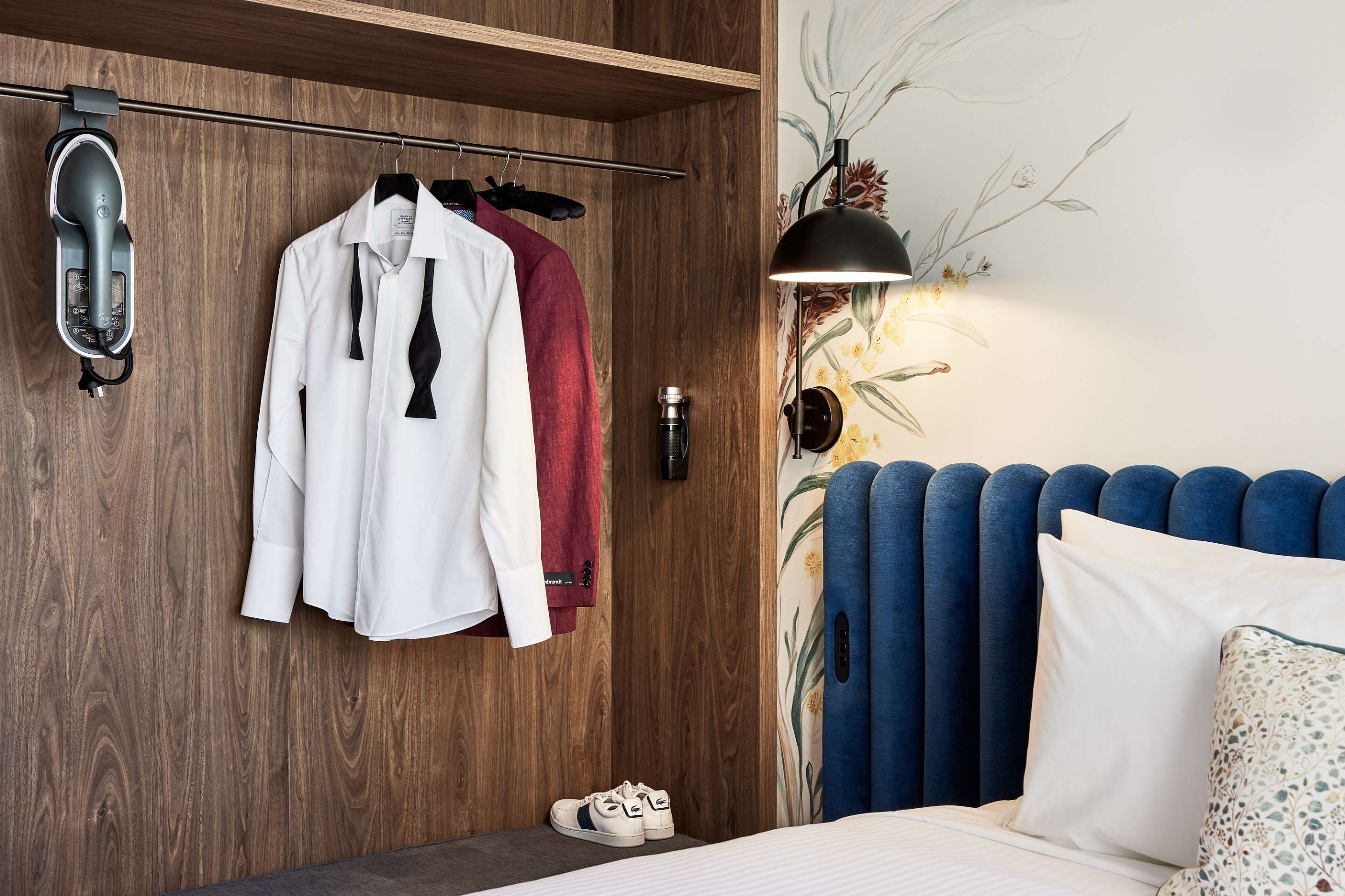 A neatly organized bedroom with a wooden wardrobe, a bed with blue headboard, and a wall-mounted lamp.