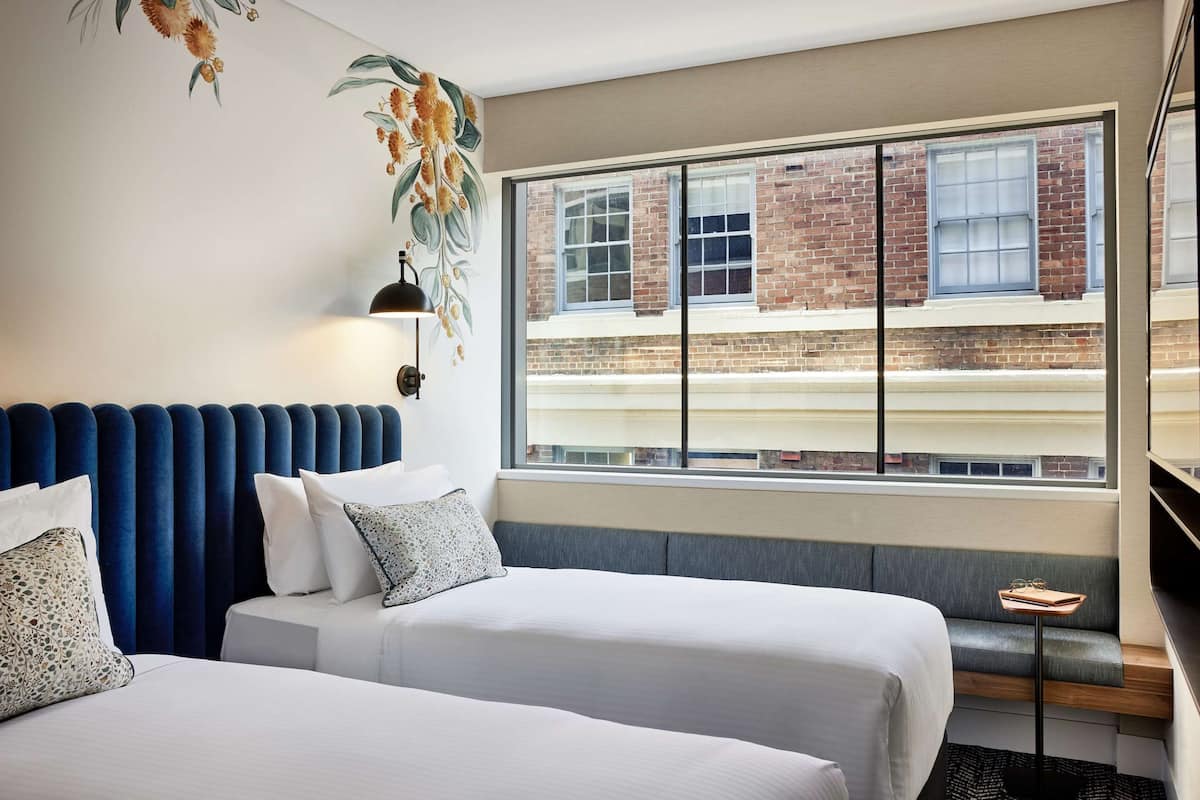 A hotel room with two beds, a large window with a view of a brick building, a blue headboard, and a small table with glasses.