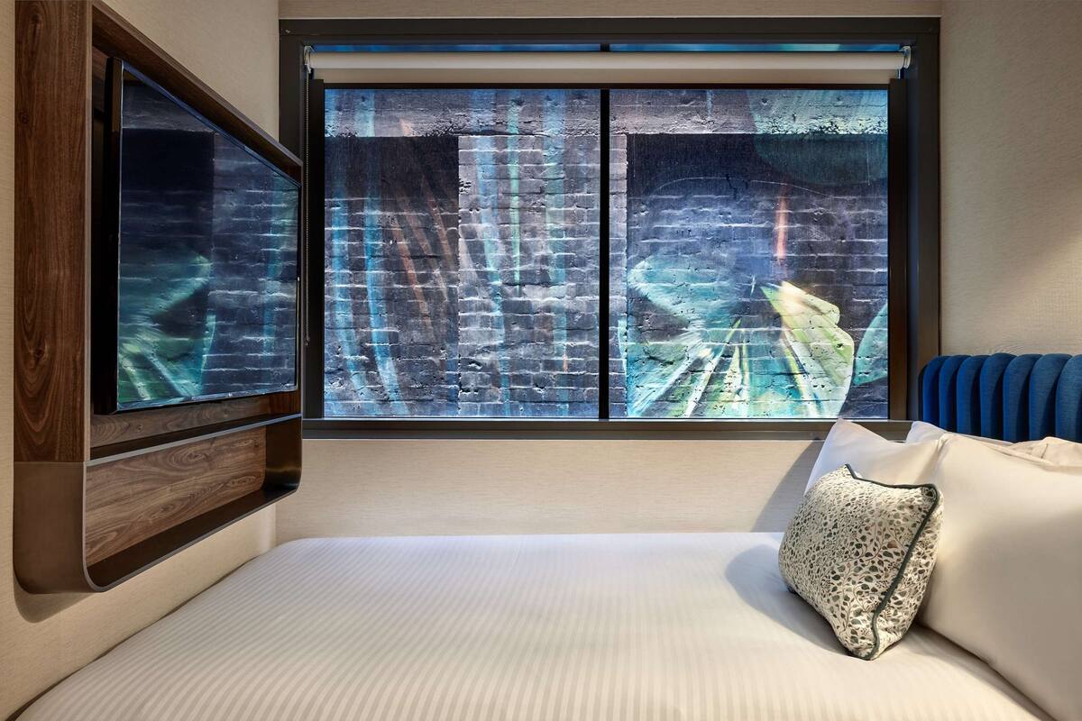 A hotel room with a bed, a large window with a view of a brick wall, and a mounted television.