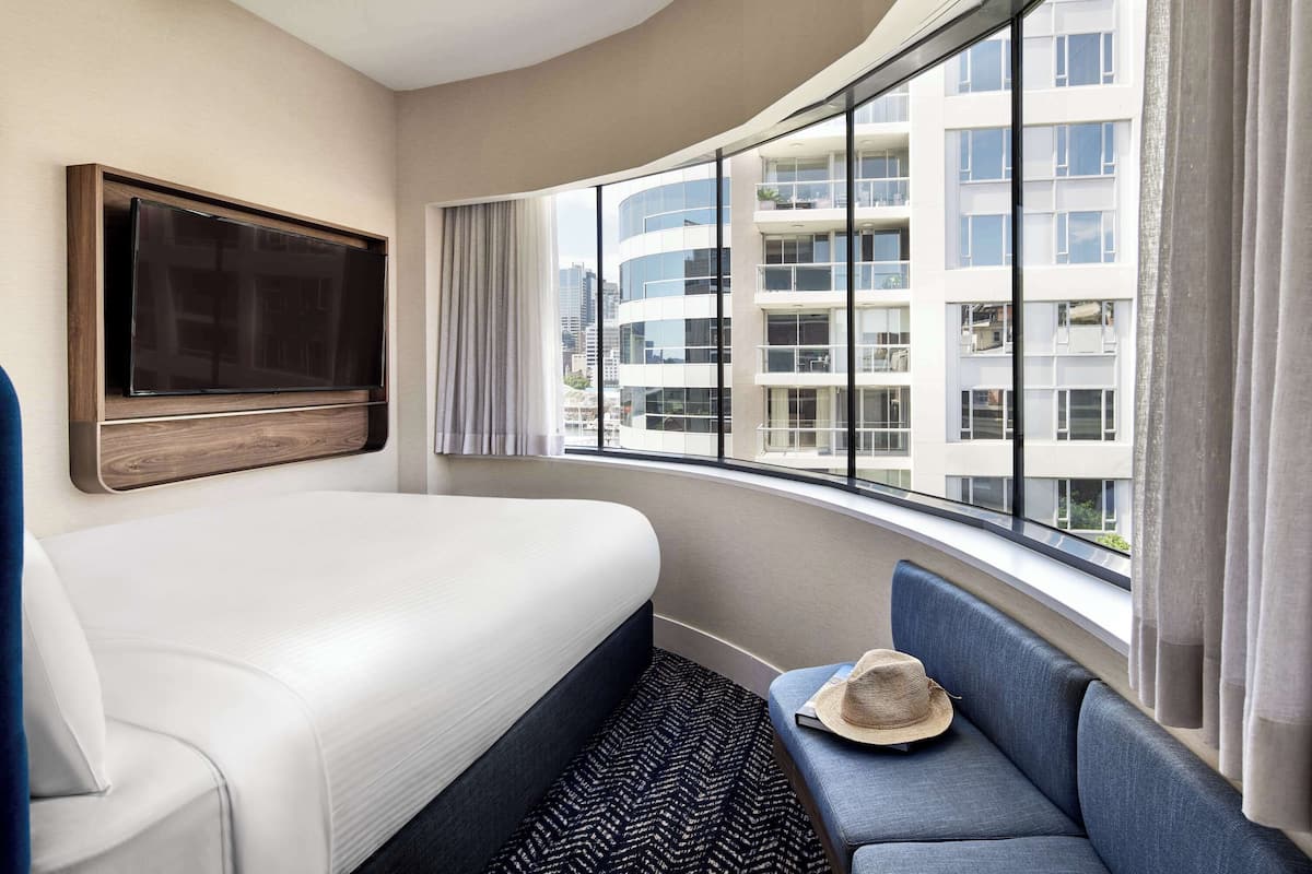 A hotel room with a large bed, a TV mounted on the wall, a seating area with a sofa and a hat, and a large window offering a view of a modern building.