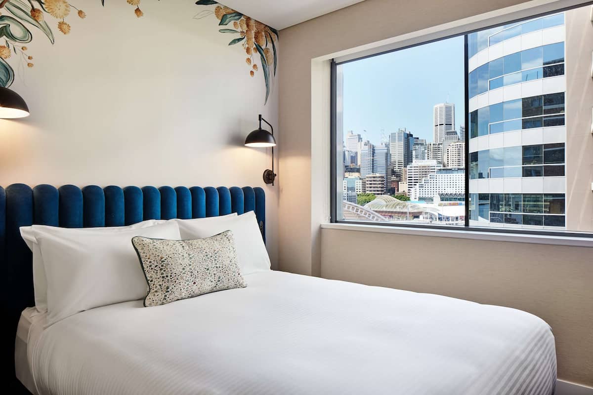 A hotel room with a large window offering a city view, a neatly made bed with white linens, and a decorative wall with floral patterns.