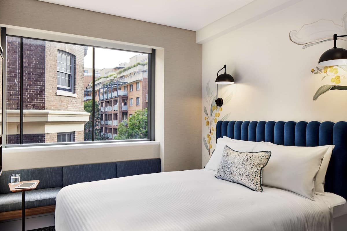 A hotel room with a large window, a bed with a blue headboard, a bedside table with a glass of water, and a wall-mounted lamp.