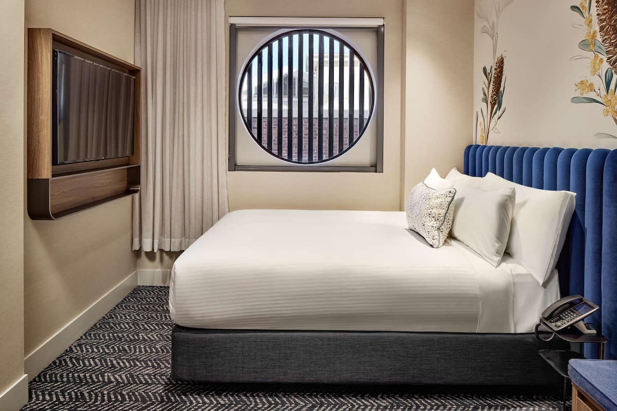 A hotel room with a large bed, a TV mounted on the wall, a window with vertical blinds, and a telephone on a side table.