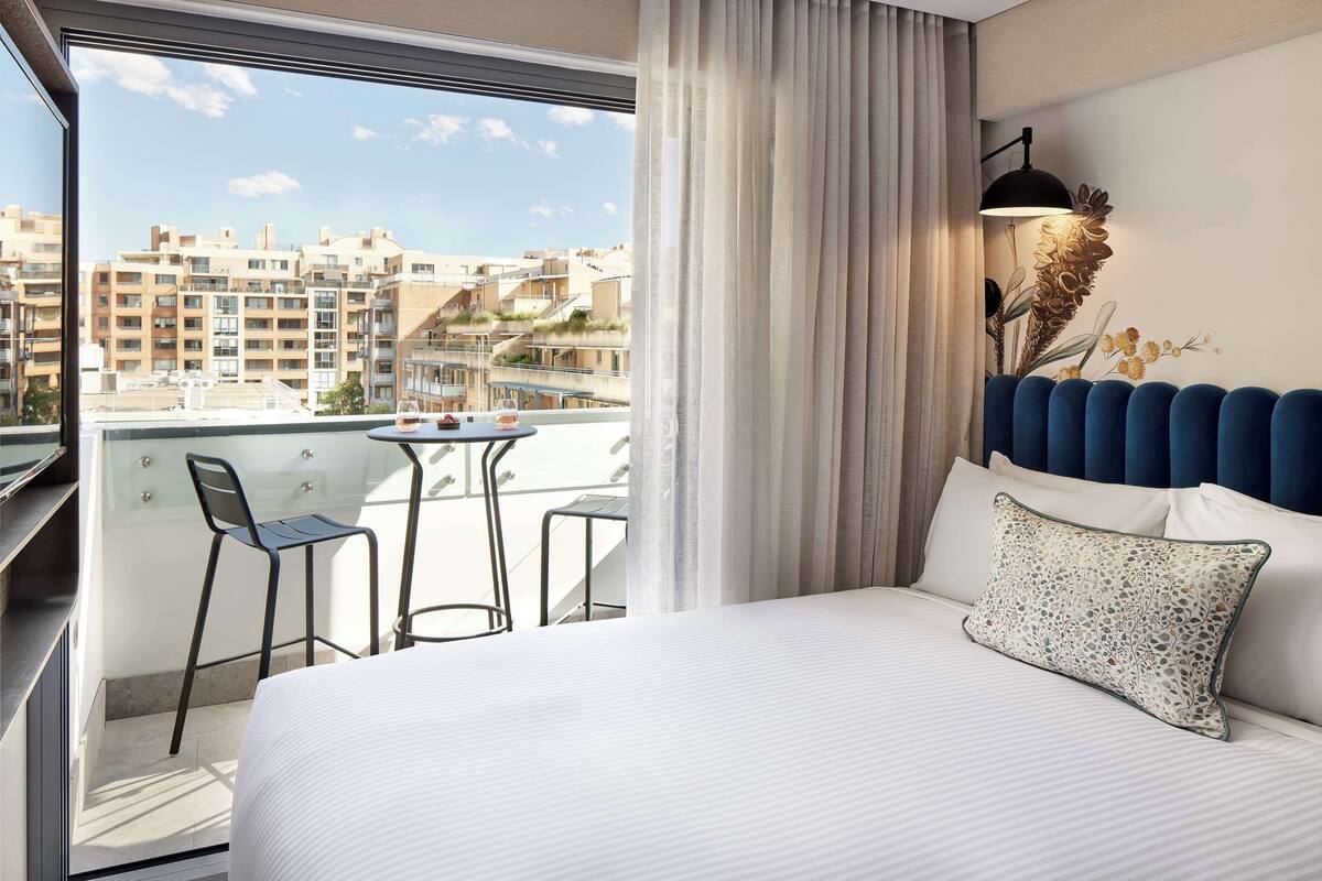 A hotel room with a large bed, a small table with two chairs, and a view of the cityscape through large windows.