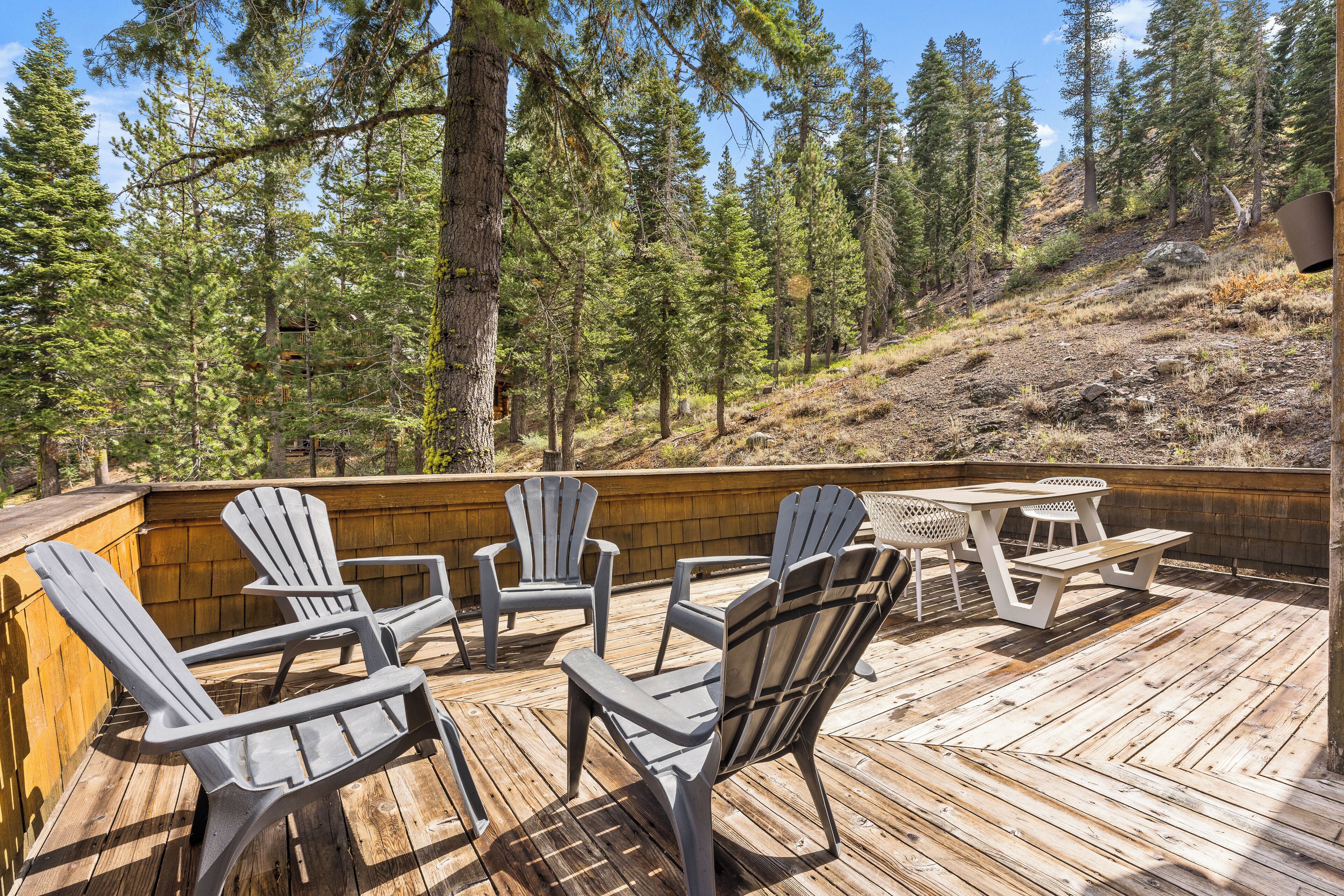 Una terraza de madera con sillas Adirondack y una mesa de picnic, rodeada por una ladera boscosa.