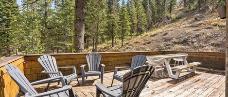 Una terraza de madera con sillas Adirondack y una mesa de picnic, rodeada por una ladera boscosa.