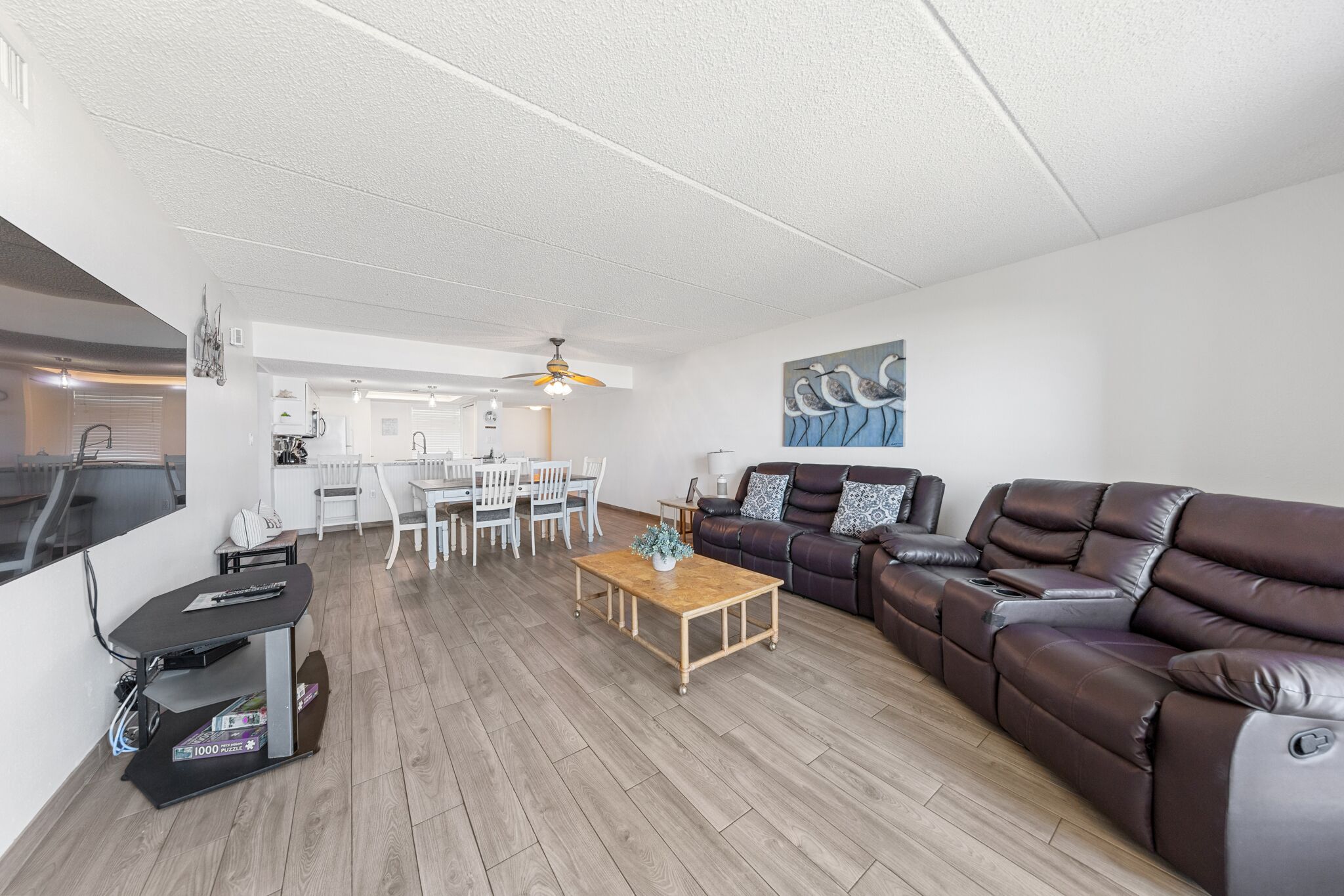 A modern living room with a brown leather sofa, a wooden coffee table, and a dining area with white chairs.