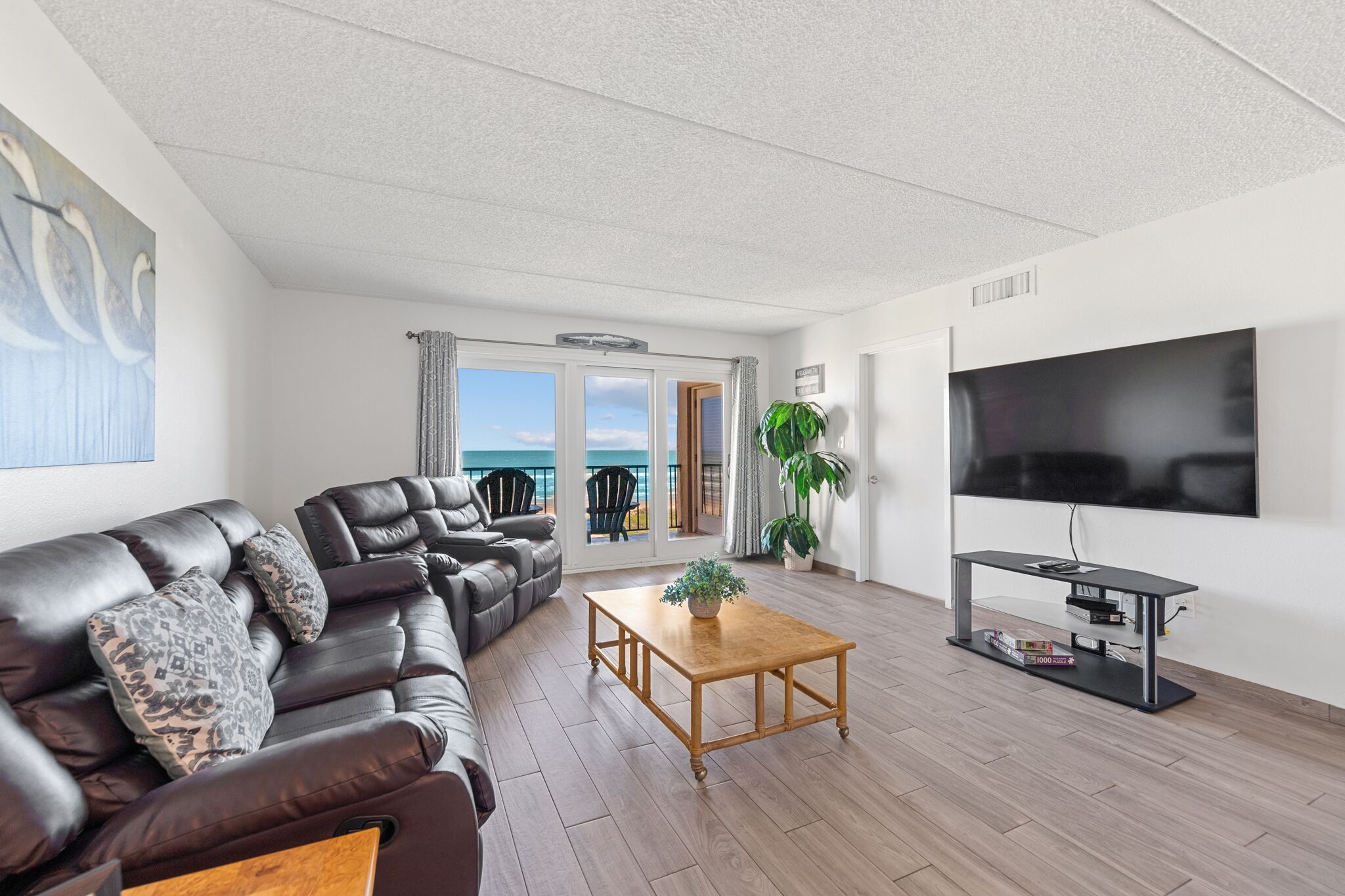 Uma sala de estar moderna com sofá de couro, mesa de centro de madeira, televisão de tela plana e varanda com vista para o mar.