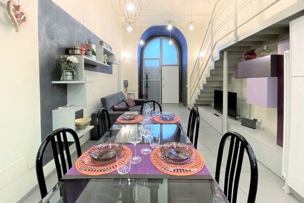 A dining area with a glass table set for six, surrounded by black chairs. There is a sofa and a staircase in the background. - LOFT 22 - Moderno a due passi da Piazza del Campo (Siena)
