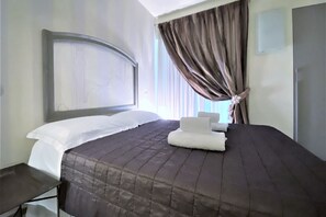 Een hotelkamer met een groot bed, hoofdeinde, nachtkastje en raam met gordijnen.