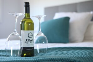A bottle of Mala Retreat wine and two wine glasses on a bed with a teal pillow.