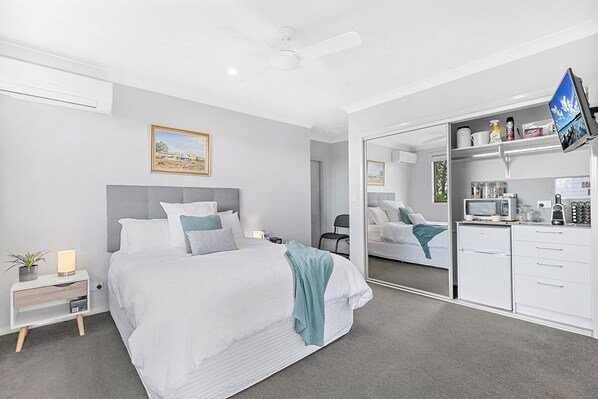 A modern bedroom with a large bed, a bedside table with a lamp, a small plant, and a mirror cabinet with a television.
