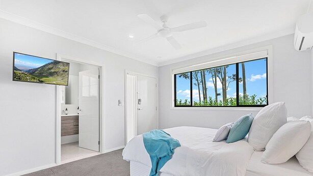 A bedroom with a bed, a ceiling fan, a large window with a view of trees, and a TV on the wall.