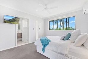 A bedroom with a bed, a ceiling fan, a large window with a view of trees, and a TV on the wall.