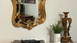 Ein Spiegel mit aufwändigem Goldrahmen, ein graues Handtuch, eine weiße Vase mit Pflanze und ein dekorativer Kerzenständer auf einer Holzkommode.