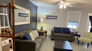 Ein Wohnzimmer mit zwei Sofas, einem Couchtisch, einem Beistelltisch und einem Deckenventilator. Zwei Wandtafeln sind angebracht, eine mit der Aufschrift 'happy place' und die andere mit 'happy home'.