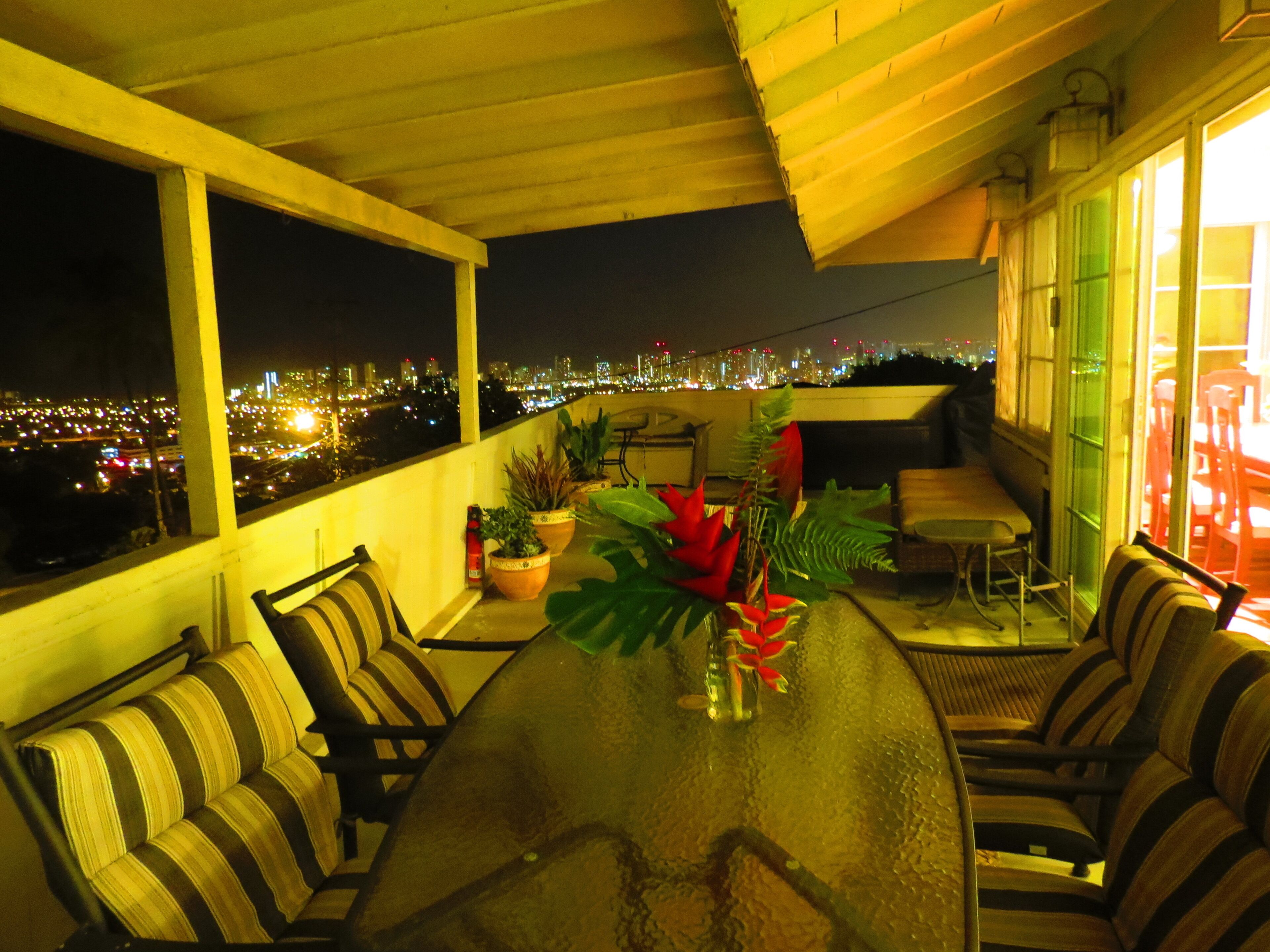 Ein Balkon mit Glastisch, gestreiften Stühlen, Topfpflanzen und Blick auf eine beleuchtete Stadtlandschaft bei Nacht.