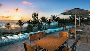 A beach resort with a pool, wooden tables and chairs, and a view of the ocean at sunset.
