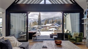 Un intérieur moderne avec une grande porte-fenêtre coulissante donnant sur un paysage montagneux enneigé. À l’intérieur, on trouve un canapé, une table basse et un coin repas avec un banc.