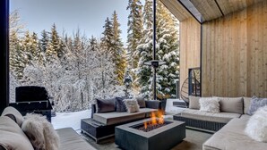 Un espace de vie extérieur moderne, doté d’un foyer, de canapés et offrant une vue sur des arbres couverts de neige.