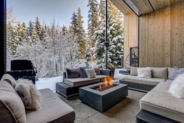 A modern outdoor living area with a fire pit, sofas, and a view of snow-covered trees.