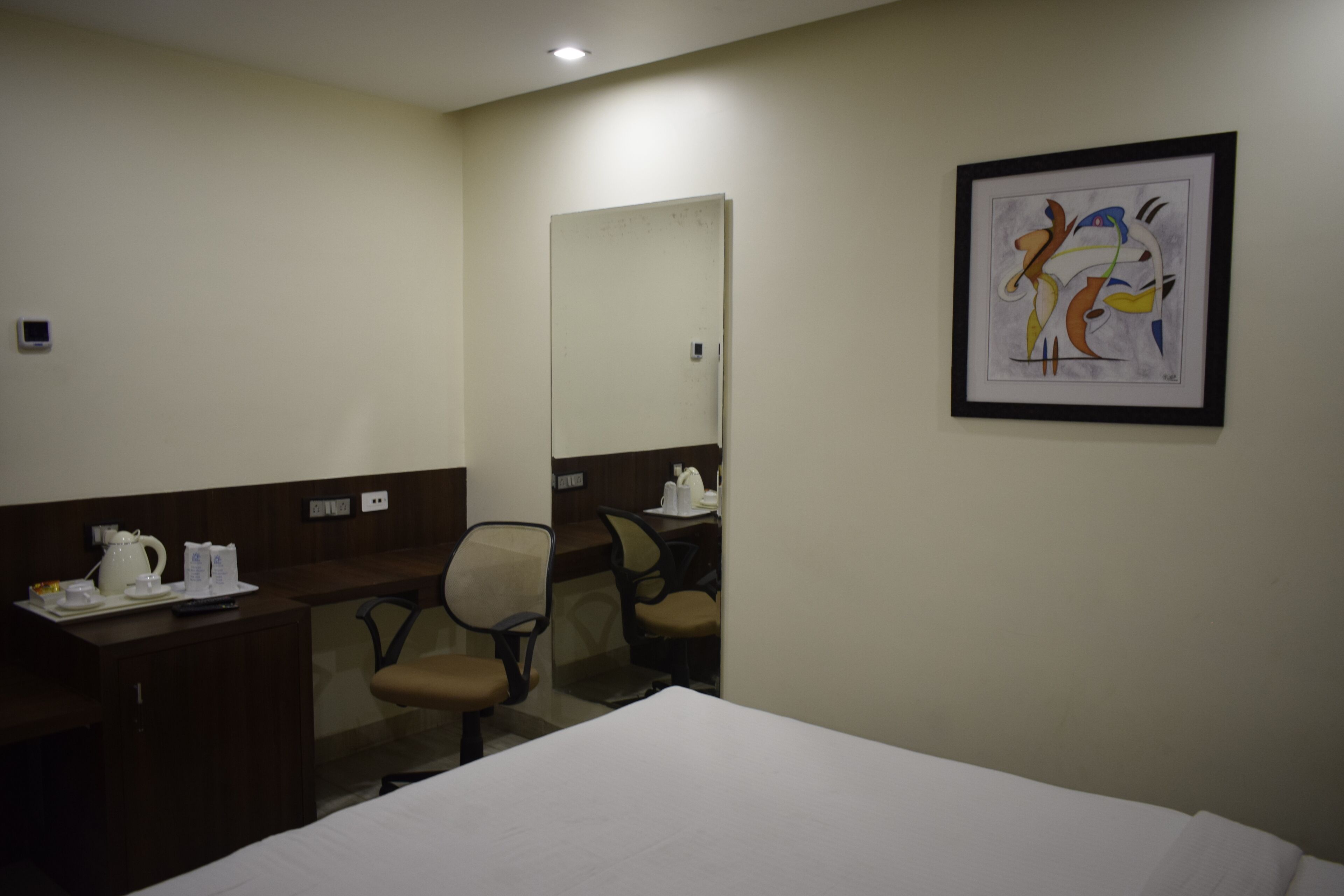 A hotel room with a bed, a desk with a chair, a framed abstract painting, and a door.