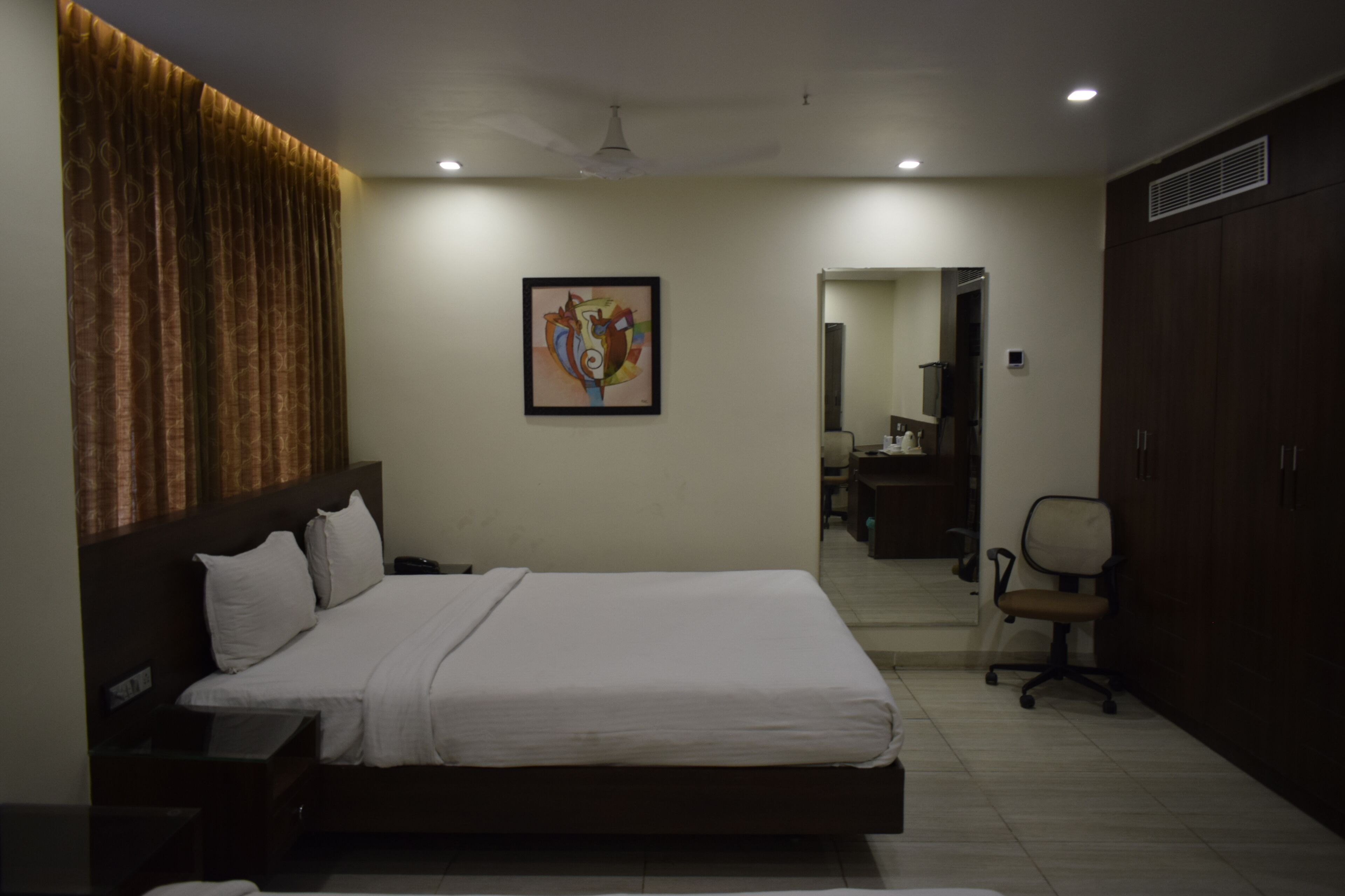 A hotel room with a large bed, a desk, a chair, a wardrobe, and a painting on the wall.