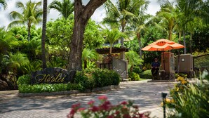 A tropical resort entrance with a stone sign, lush greenery, and a seating area under an umbrella. - I.P.M PRESENTS: HOOLEI 93-1: 3 BEDROOMS + OFFICE. FLEXIBLE CANCELATION POLICY (Kihei)