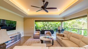 A spacious living room with a ceiling fan, a flat-screen TV, and a large window overlooking a lush garden. - I.P.M PRESENTS: HOOLEI 93-1: 3 BEDROOMS + OFFICE. FLEXIBLE CANCELATION POLICY (Kihei)