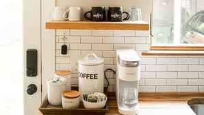 A kitchen with white tiles, wooden shelves, a coffee maker, coffee cups, sugar containers, and a plant. - The Cozy Cabernet - short walk to Main St! (Fredericksburg)