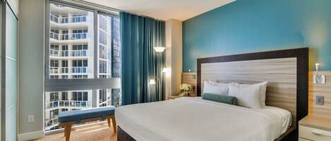 A hotel room with a large bed, a bench, a nightstand, a lamp, and a view of a high-rise building.