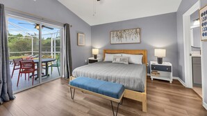 Une chambre avec un lit, un banc, un ventilateur de plafond, une table de chevet, une lampe et une vue sur un espace extérieur aménagé avec des chaises et une table, accessible par une porte-fenêtre coulissante.